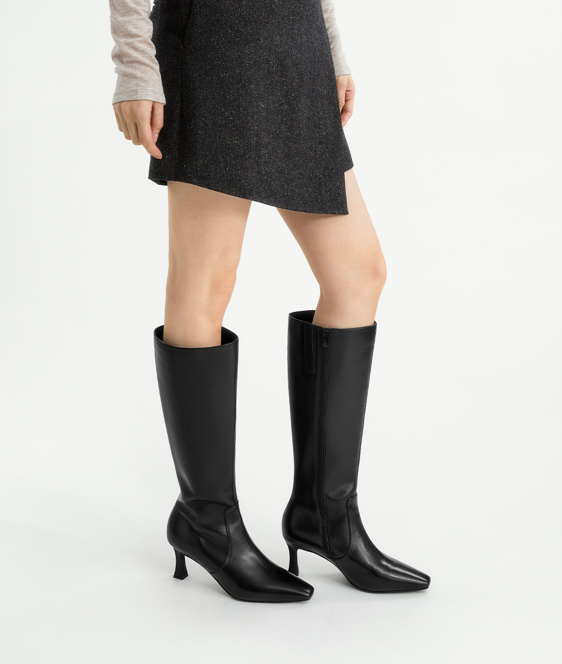 A model wearing the black pointed-toe tall riding boots with a distinctive flared heel